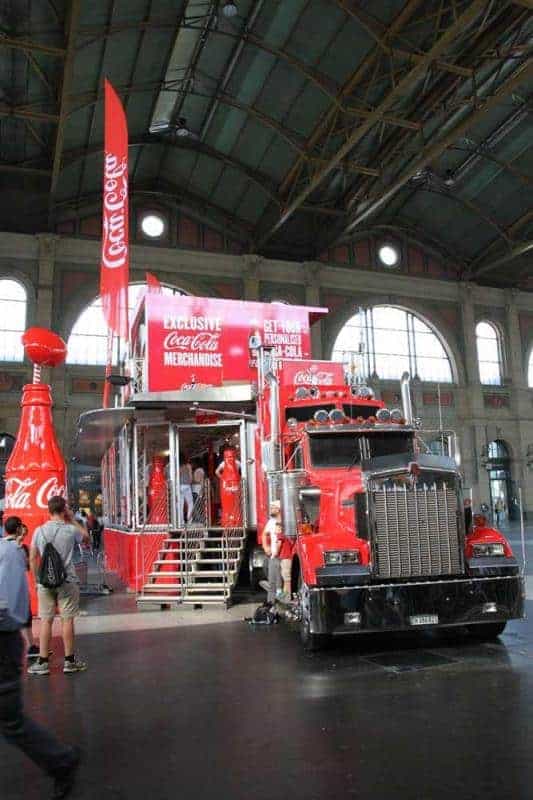 100th anniversary of the Coca-Cola glass bottle 3 Czerwony ciężarówka Coca-Cola z naczepą przekształconą w stoisko sprzedażowe z produktami Coca-Cola, stojąca w dużej hali.