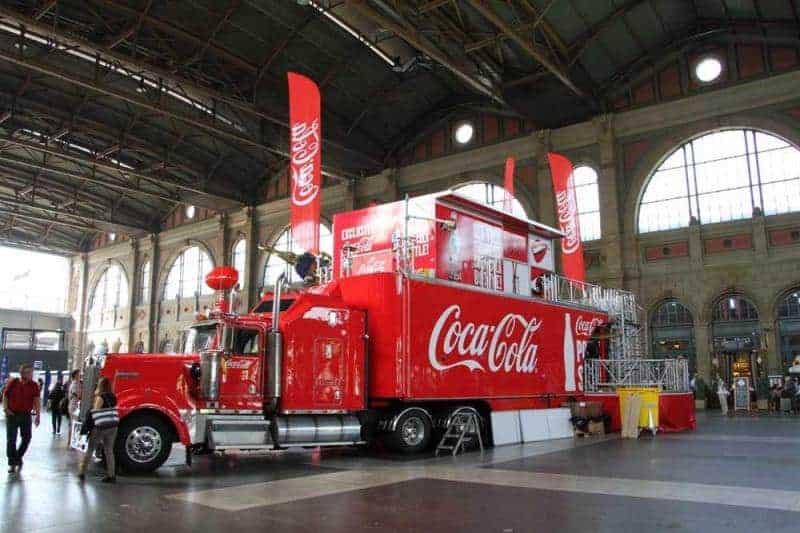 100th anniversary of the Coca-Cola glass bottle 4 Czerwony ciężarówka Coca-Cola w hali dworca z dużymi flagami Coca-Cola i dużymi oknami w tle.