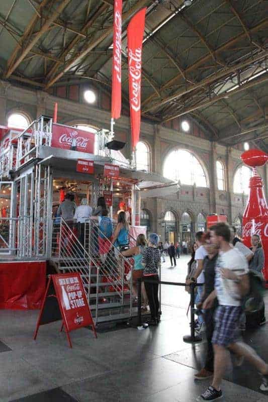 100th anniversary of the Coca-Cola glass bottle 2 Stoisko Coca-Coli z ludźmi w hali dworca kolejowego.
