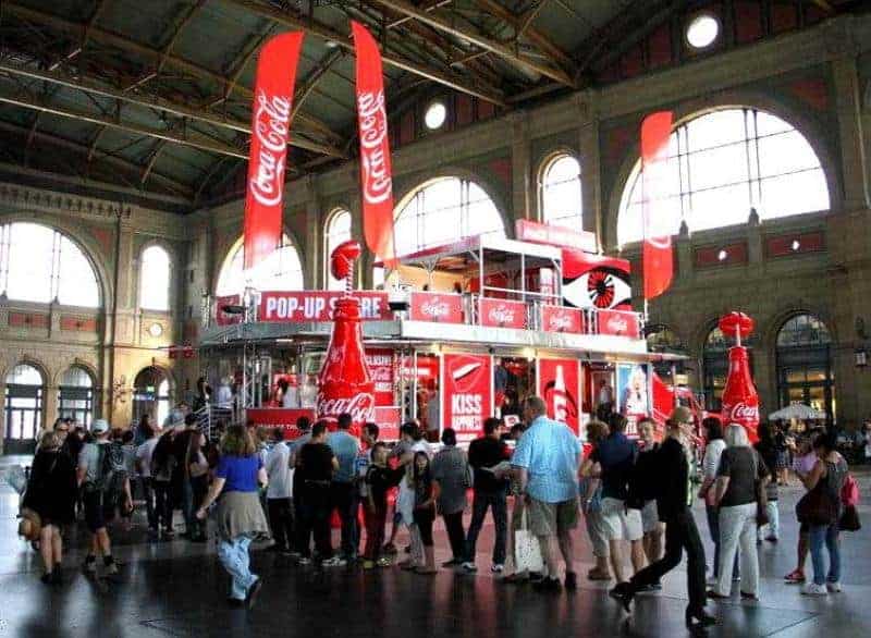 oryg_064490685_1129949550544944_1465385417682452480_o Coca-Cola pop-up store inside a large building with arched windows, surrounded by a crowd of people.