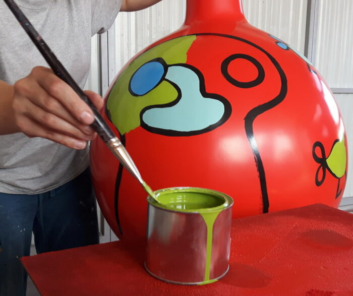 Person painting a large red vase with abstract designs using green paint.