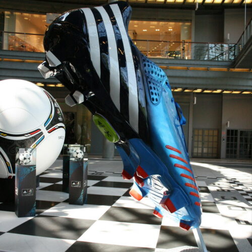 Giant colorful soccer cleat sculpture on a checkered floor with a large soccer ball in the background, situated in an indoor space with modern architecture.
