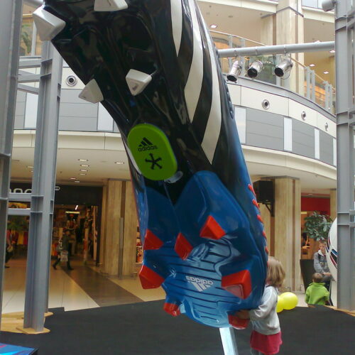 A giant model of an Adidas football boot displayed in a public indoor area, with a small child standing near the boot holding yellow balloons.