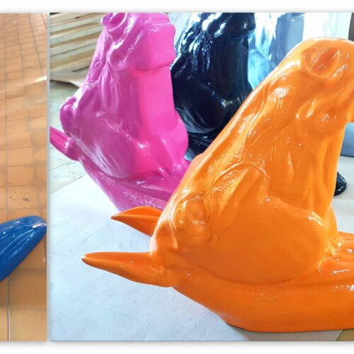 Colorful sculptures of dogs lying down, featuring glossy finishes in various colors including blue, orange, pink, white, black, and green, are arranged in rows on a tiled floor.