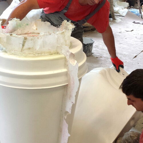 Men working on a large cylindrical mold in a workshop, wearing red shirts, gloves, and overalls.