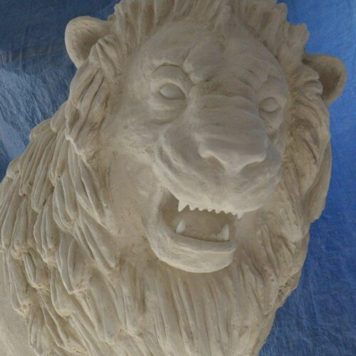 Close-up view of a detailed lion statue with an open mouth, showing teeth against a blue background.