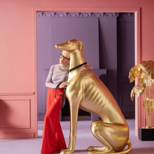 An elegantly dressed woman standing beside and leaning on a large golden dog statue in a room with pink and purple walls.