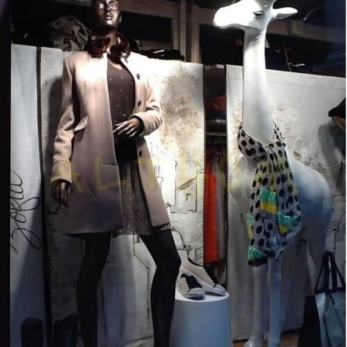 Store window display featuring a mannequin dressed in a beige coat and dotted dress, and a white giraffe model with a colorful scarf.