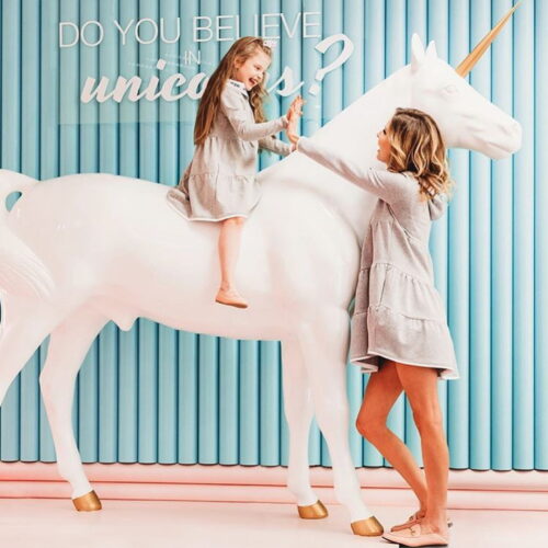 A woman and a child, both wearing matching grey dresses, interact happily with a large white unicorn statue in front of a blue wall with the text
