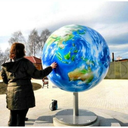 A person in a winter coat touches a large outdoor globe sculpture mounted on a stand, with a park and trees in the background.