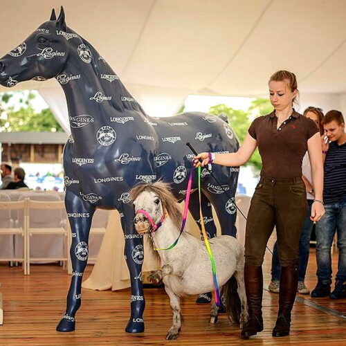 A woman leads a small pony on a colorful rope leash, walking past a dark blue model horse covered in Longines logos.