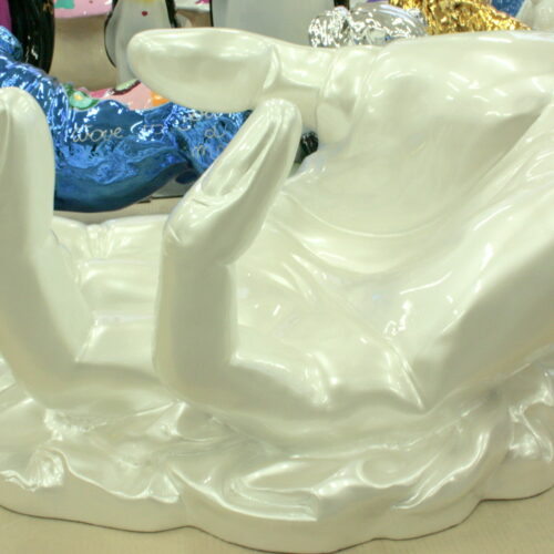White sculpture of a hand with fingers extended upward, displayed on a reflective surface with other colorful sculptures in the background.