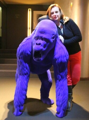 Woman posing with a purple gorilla statue indoors.