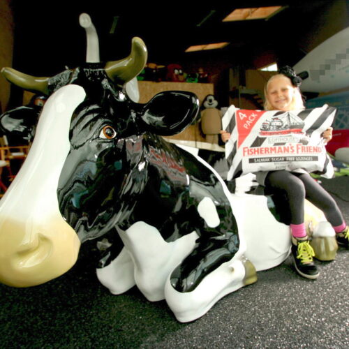 Child sitting on a large statue of a black and white cow, holding a giant Fisherman's Friend lozenge pack.