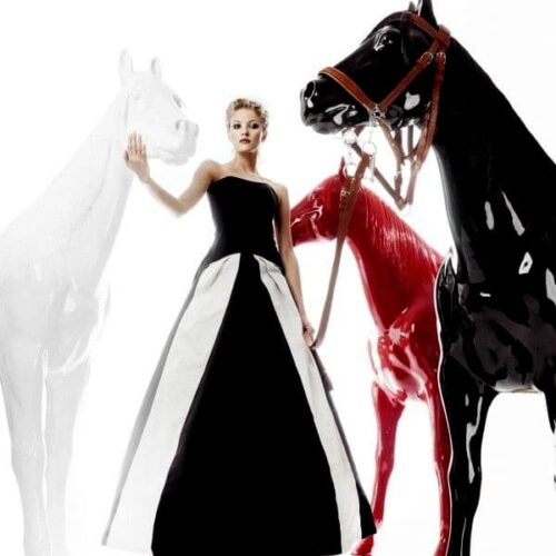 Woman in a black and white gown standing among three large, decorative horse figures in black, white, and red colors.