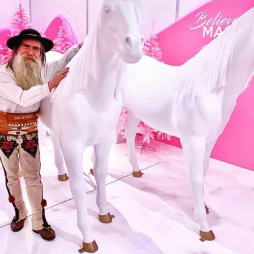 A man in traditional attire stands next to two white horse statues in a pink-themed room with the phrase