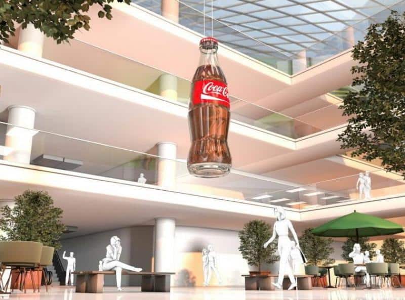 oryg_064490685_1129949550544944_1465385417682452480_o Large Coca-Cola bottle suspended in a multi-level indoor space with people and greenery.