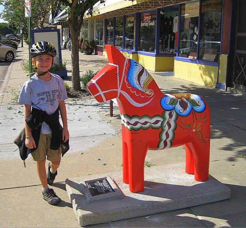 Artistically painted horses from Dalarna 2 Chłopiec w kasku na rower stojący obok kolorowej rzeźby konia na chodniku wzdłuż ulicy z witrynami sklepów.