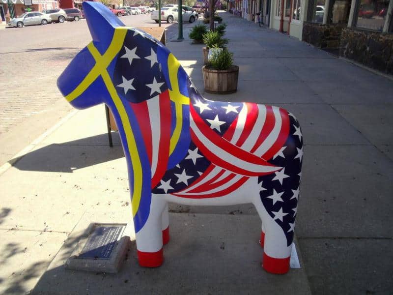 oryg_064490685_1129949550544944_1465385417682452480_o Colorful painted Dala horse statue on a city sidewalk, featuring a design combining American flag patterns with elements of the Swedish flag.