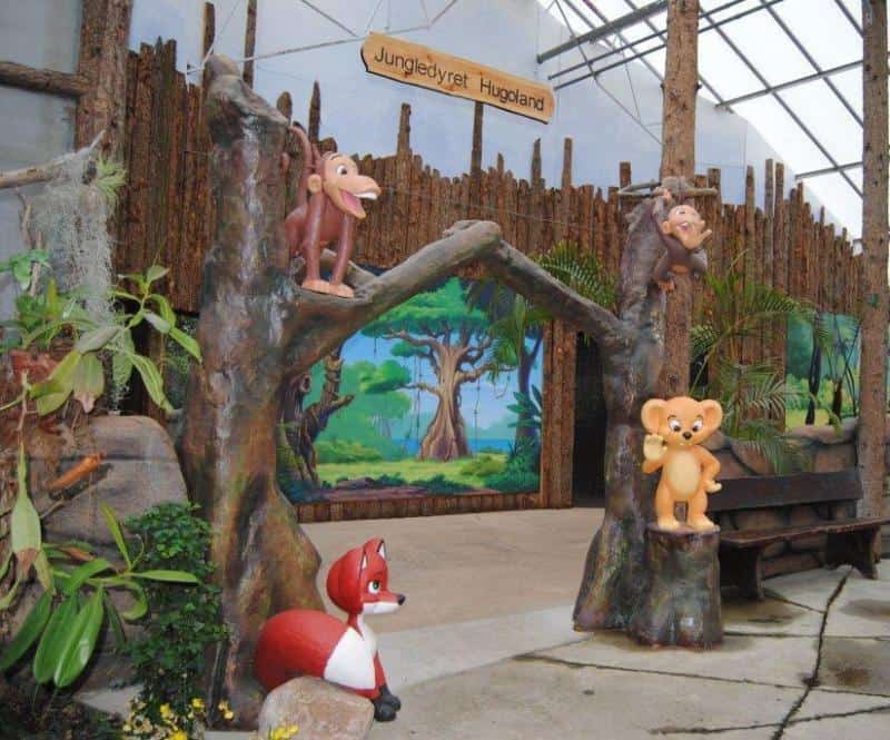 oryg_064490685_1129949550544944_1465385417682452480_o Wooden archway with cartoon animal statues, including a monkey, fox, and lion cub, under a sign reading "Jungledyret Hugoland."