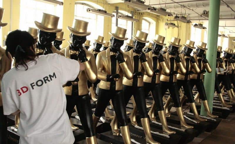Large Johnnie Walker Figures 4 Rzędy manekinów ubrane we złote garnitury i cylindry, osoba w białej koszulce z napisem "3D FORM" pracuje przy jednym z nich.