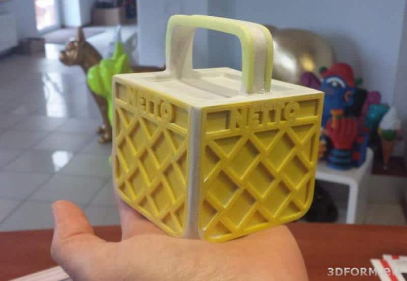 NETTO 3D LOGO 4 Mały model żółtej skrzynki z napisem "NETTO", trzymany na dłoni.