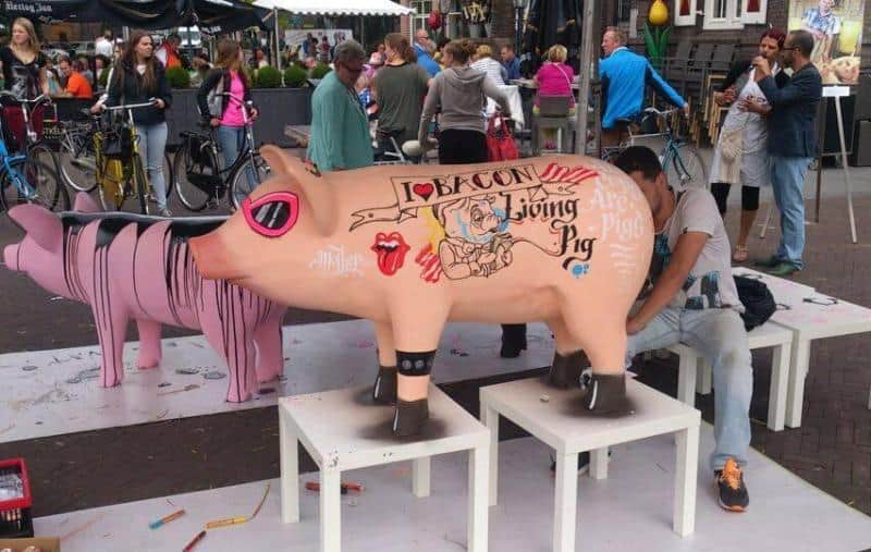 oryg_064490685_1129949550544944_1465385417682452480_o People in an outdoor plaza painting cartoonish pig sculptures. One pig sculpture is painted with a tattoo-style design featuring the words "I Love Bacon" and "Flying Pig" along with various drawings.