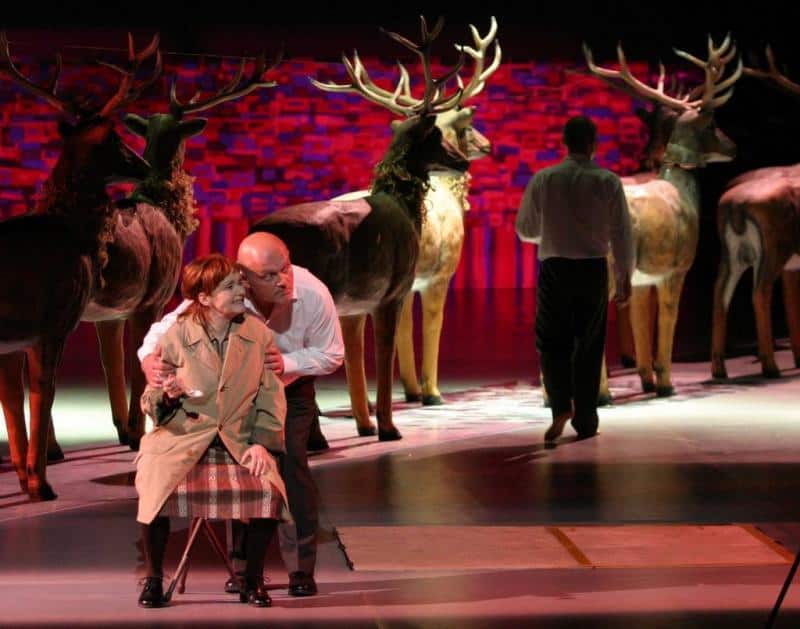 oryg_064490685_1129949550544944_1465385417682452480_o Stage performance featuring two actors, one sitting and the other kneeling, with a backdrop of reindeer and a vibrant street art wall.