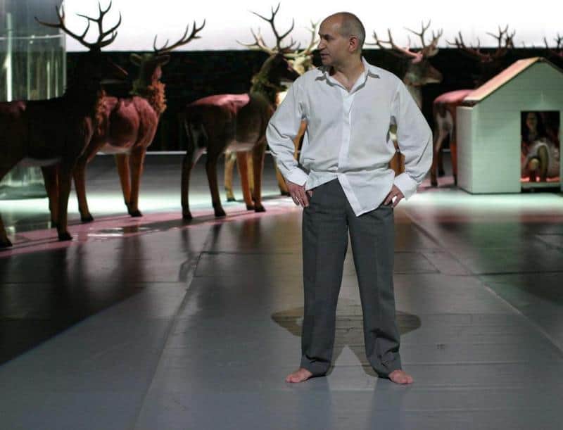 Stage design elements for the Dramatic Theatre of the Capital City of Warsaw 1 Mężczyzna w białej koszuli i szarych spodniach stoi boso na scenie, w tle stoją wypchane jelenie.