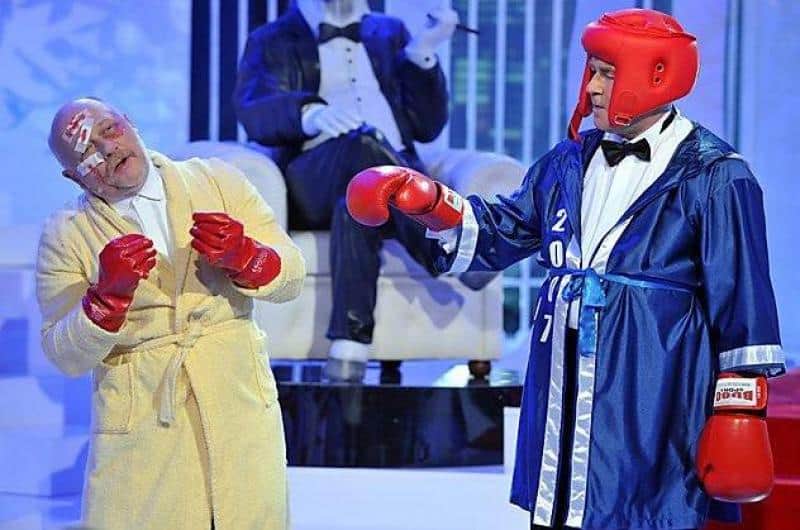 TVP2 - Set Design Elements 2 Dwóch mężczyzn ubranych w stroje bokserskie i rękawice bokserskie podczas występu scenicznego, jeden w szlafroku, a drugi w niebieskim szlafroku bokserskim i kasku ochronnym.
