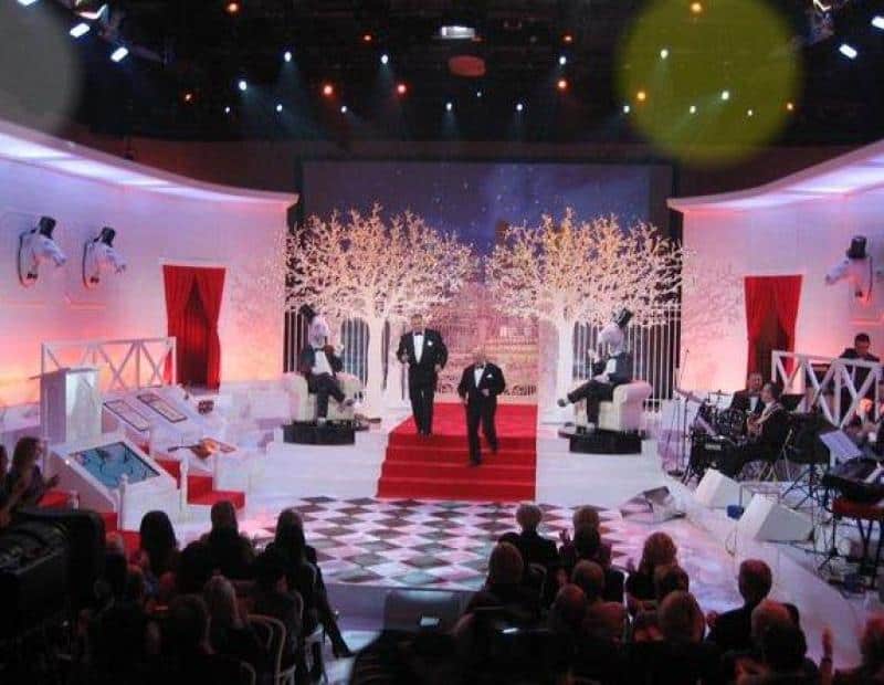 oryg_064490685_1129949550544944_1465385417682452480_o Night show stage with red carpet, two men in tuxedos, lit white trees, seated penguin mannequins, audience in foreground, and musicians to the right