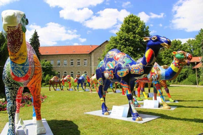 oryg_064490685_1129949550544944_1465385417682452480_o Colorful camel sculptures displayed in an outdoor park with a red-roofed building and trees in the background.