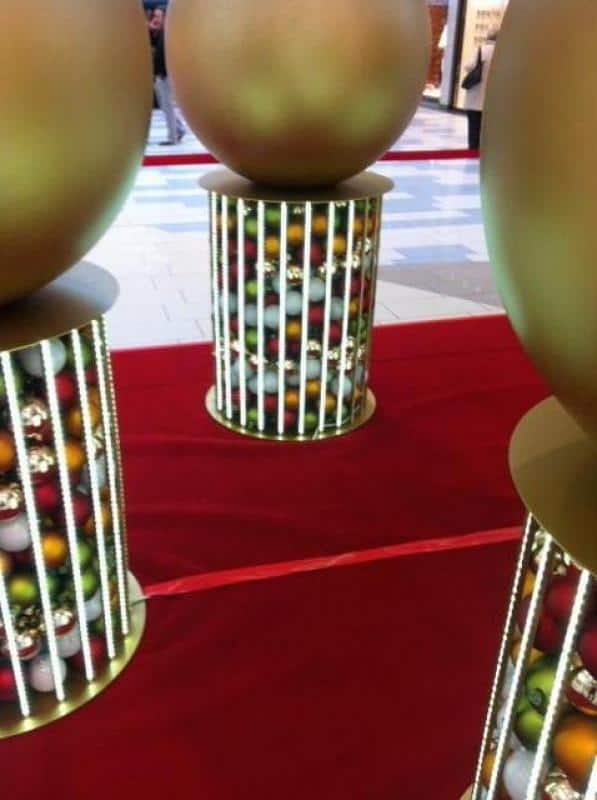 Illuminated Christmas baubles 3 Duże złote kule na cylindrycznych stojakach w galerii handlowej.