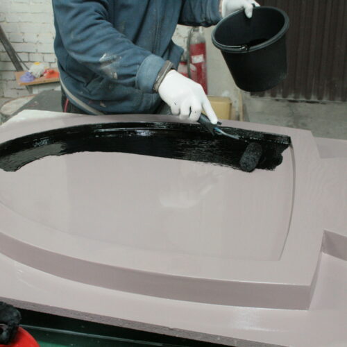 A person wearing gloves paints a large mold with black paint using a roller in a workshop.