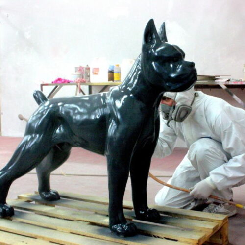 A person wearing a protective suit and mask is spray-painting a large, shiny black statue of a dog standing on a wooden pallet.