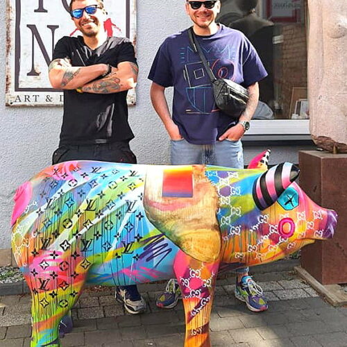 Two men stand next to a colorful, multicolored pig statue with various brand logos, outside a shop.