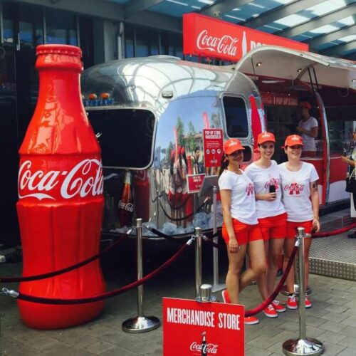 Large Coca-Cola bottle model next to a silver airstream trailer with Coca-Cola signage; three women in Coca-Cola branded outfits are standing in front of the trailer, which is labeled as a