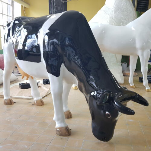 A black-and-white cow statue with a glossy finish stands on a tiled floor in an indoor setting. The cow is in a grazing position with its head bent down.
