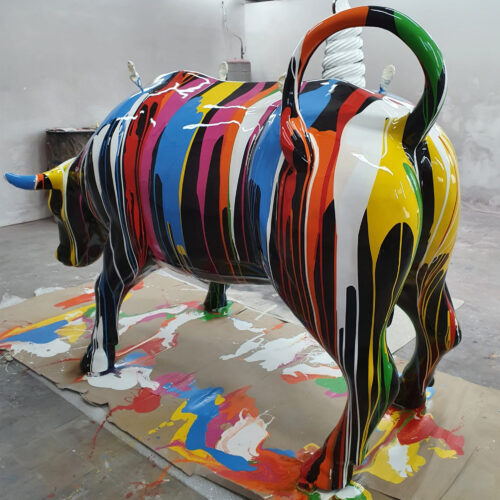 A life-sized fiberglass bull statue painted with vibrant, multicolored dripping stripes standing on a cardboard surface covered with splatters of paint.