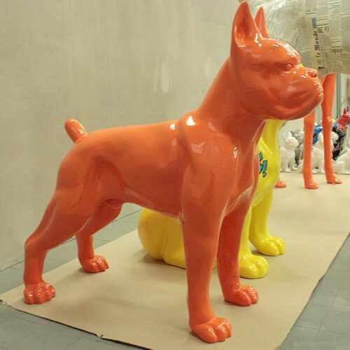 Large orange dog sculpture with a shiny surface standing on a beige platform, with partial views of yellow and white dog sculptures in the background.