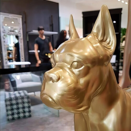 A golden statue of a dog with focused detailing, positioned indoors with a blurred background reflecting people and furniture.