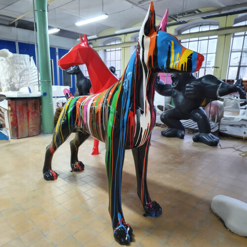 Colorful life-sized sculpture of a dog with multicolored paint drips, inside an art studio with other large sculptures in the background.