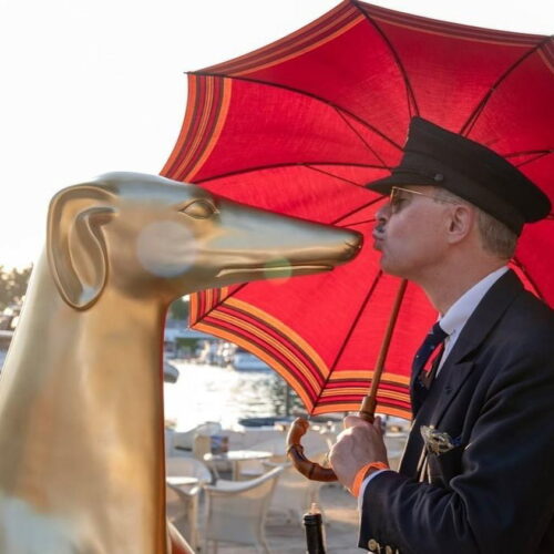 A man holding a red umbrella kisses a golden greyhound statue outdoors near a waterfront.