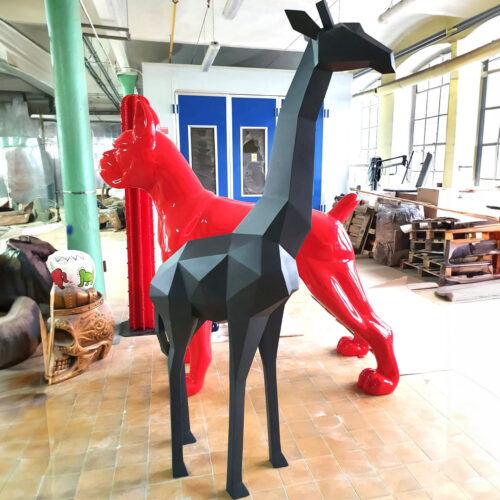 Geometric black giraffe sculpture in front of a glossy red dog sculpture, in a workshop setting with tools and materials in the background.