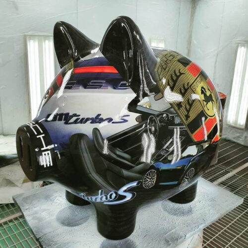A black piggy bank with a glossy finish, featuring various automotive-themed graphics such as a car interior, the Porsche logo, and the word