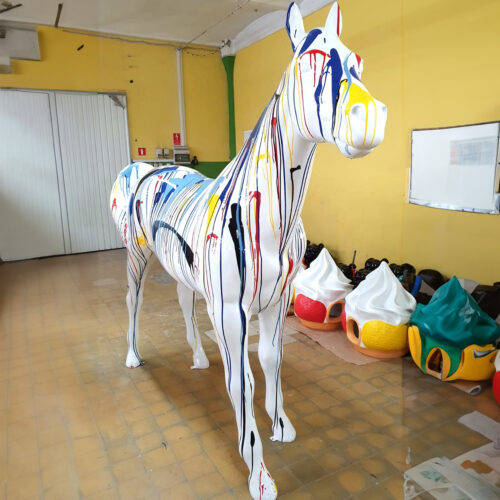 A large white horse sculpture with colorful paint drips, standing in a yellow room with various decorative objects in the background.