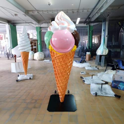 Large plastic ice cream cone sculptures in a workshop, with a prominent sculpture featuring scoops of pink, green, and brown ice cream topped with white whipped cream on a waffle cone.