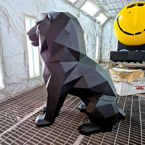 Geometric black lion sculpture inside a workshop with a yellow smiley face sculpture in the background.