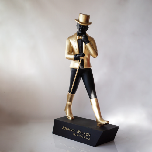 Statue of a man in black and gold clothing, holding a walking cane, standing on a black base with the inscription