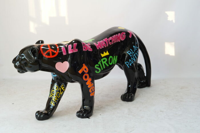 Black panther statue adorned with colorful words and symbols, including "POWER," "BLACK PANTHER," "I'LL BE WATCHING YOU," a pink heart, and a red peace sign.